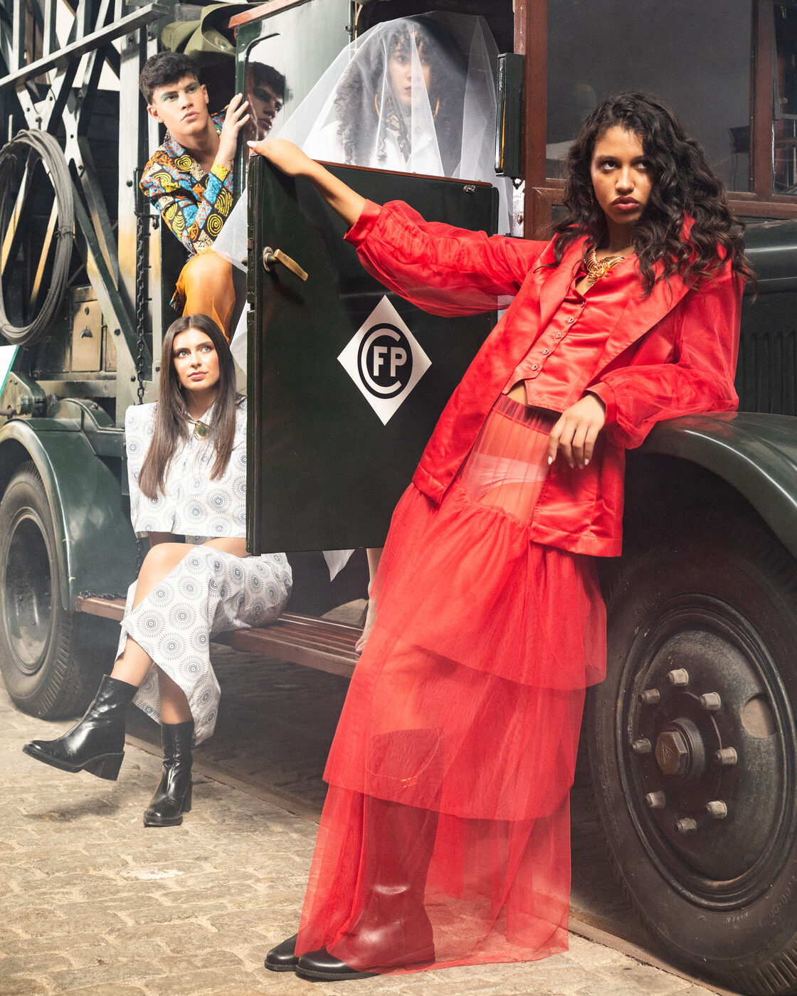 Modelo em look Flame Scarlet com saia de tule apoiada na porta do camião; ao lado, conjunto estampado e véu de tule