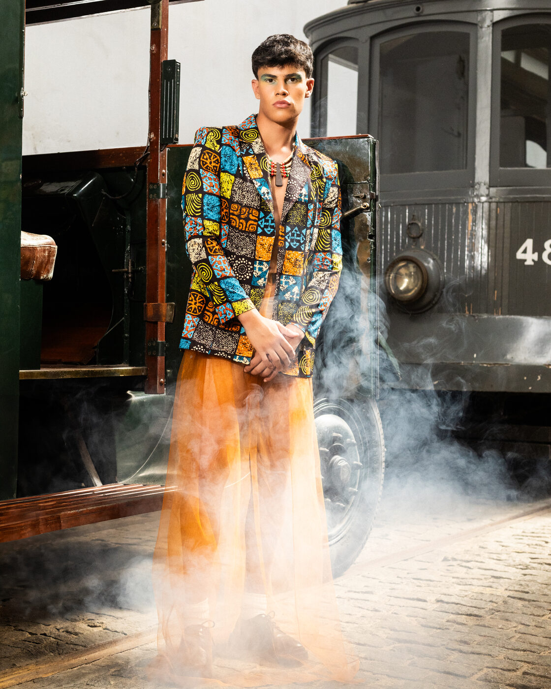 Modelo em blazer estampado e saia de tule Orangeade em plano geral, diante de um elétrico antigo, entre fumo