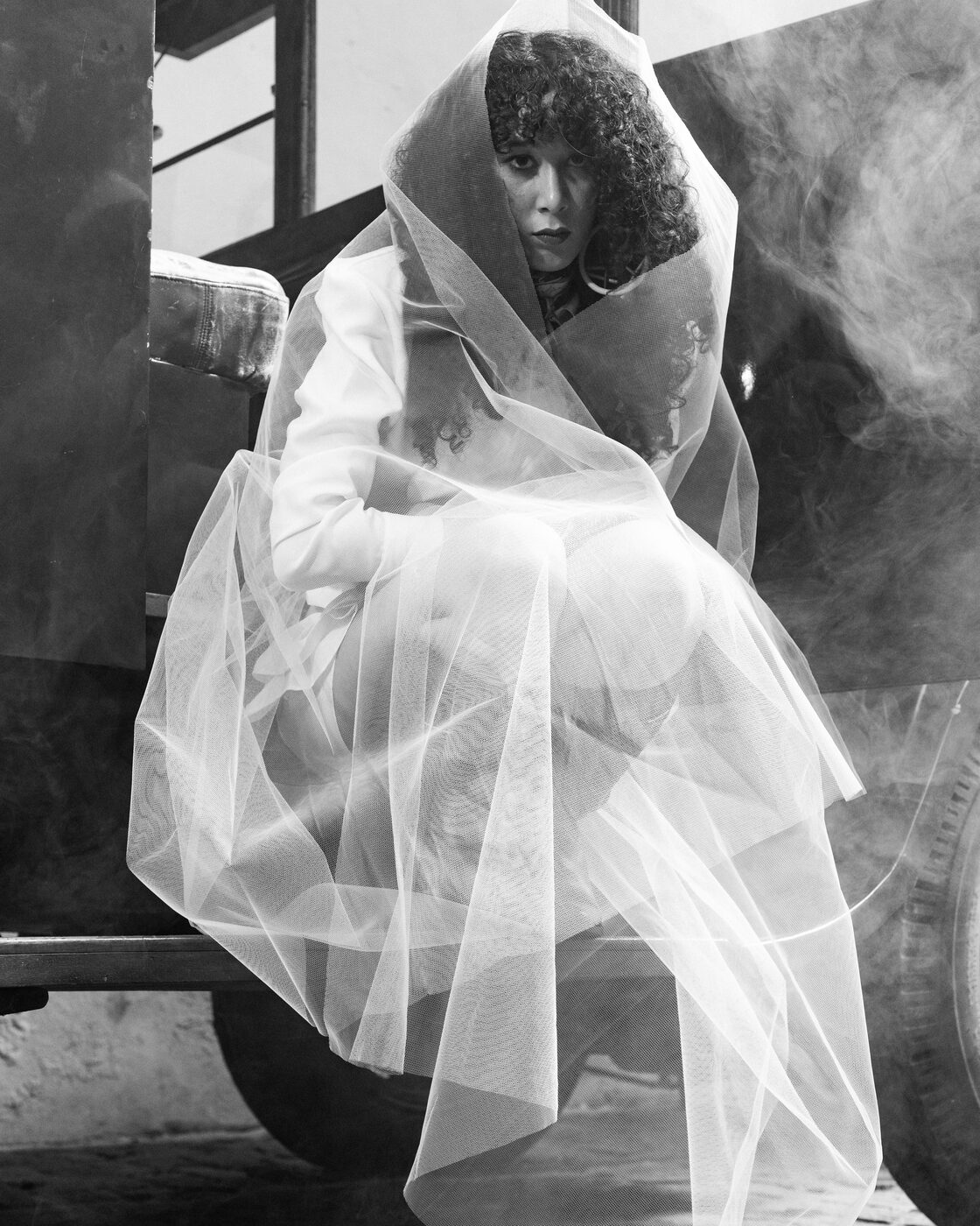Fotografia a preto e branco: modelo envolta em véu de tule sentada no estribo do camião, entre fumo