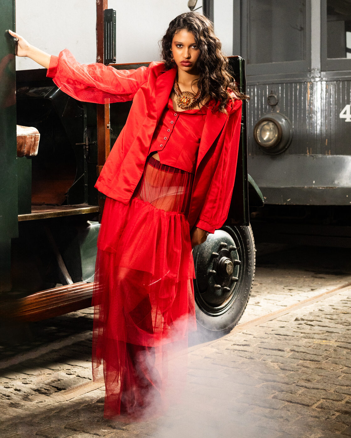 Modelo em look Flame Scarlet completo — casaco, corpete e saia de tule em camadas — junto à roda do camião, entre fumo